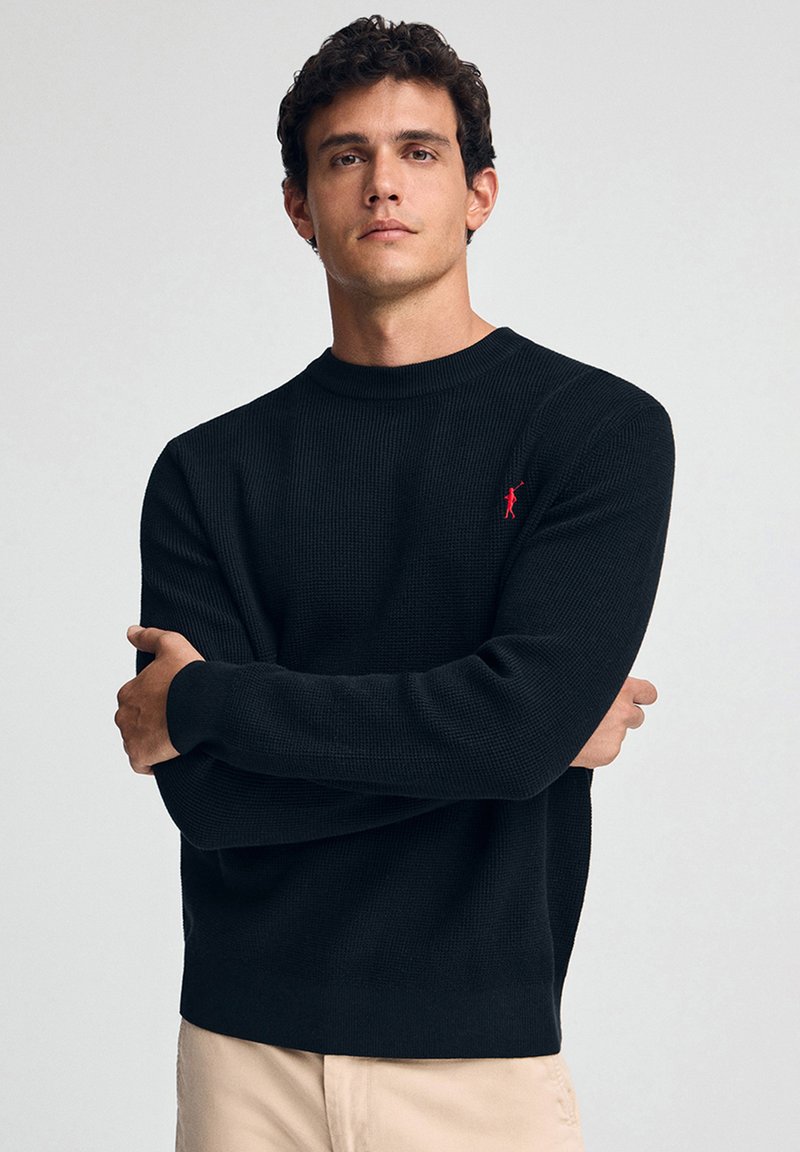 Schwarzer, gerippter Strickpullover mit Rundhalsausschnitt und langen Ärmeln. Verfügt über ein kleines rotes Logo auf der Brust. Eng anliegendes Design.