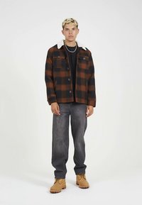 Καρό μπουφάν σε καφέ και μαύρο χρώμα με λευκό γιακά από συνθετικό μαλλί, κουμπώνει με κουμπιά, έχει δύο τσέπες στο στήθος, συνδυασμένο με γκρί τζιν και καφέ μπότες.