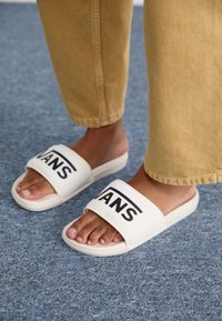 Bijele sandale na klizanju s širokim remenom koji sadrži crni "VANS" logo. Potplat je ravna, a dizajn je minimalistički. Uparene s bež skraćenim hlačama.