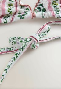 Rózsaszín és zöld virágmintás bikini felső hullámos szegéllyel, bézs háttéren, megkötős szalagos pánttal.