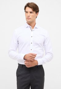 Camicia bianca con maniche lunghe, caratterizzata da un colletto a contrasto con motivo e bottoni marroni. Tessuto liscio, vestibilità sartoriale, indossata con pantaloni scuri.