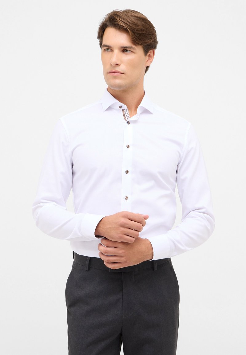 Camicia bianca con maniche lunghe, caratterizzata da un colletto a contrasto con motivo e bottoni marroni. Tessuto liscio, vestibilità sartoriale, indossata con pantaloni scuri.