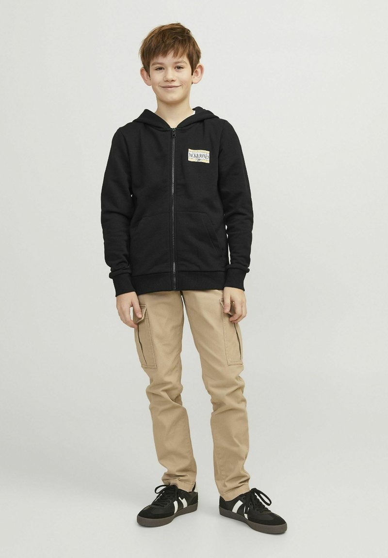 Haut de sweat à zip noir avec poches avant et patch logo, pantalon cargo beige avec poches latérales, et baskets noires avec rayures blanches.