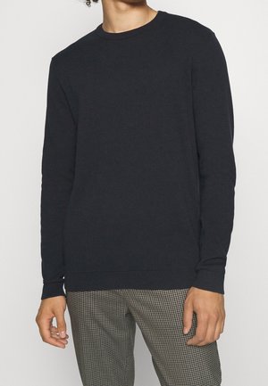 Dunkelblauer Rundhalsausschnitt-Pullover, mit gerippten Bündchen und Saum, aus weichem Material gefertigt. Kombiniert mit karierter Hose. Minimalistisches Design und Passform.