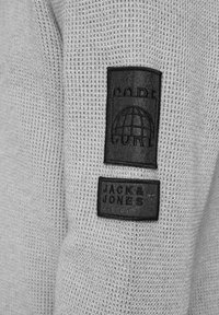 Suéter gris de punto con dos etiquetas de tela negras con texto en relieve: "CODE" y "JACK & JONES 1984." Textura ligera.
