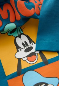 Turkis t-shirt med et grafisk print af Goofys ansigt i sort og hvidt mod en gul og orange baggrund med legende tekst.