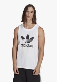 Balta medvilninė liemenė, kurios priekyje yra didelis juodas Adidas logotipas ir trefoil dizainas. Apvalus iškirpimas ir be rankovių.