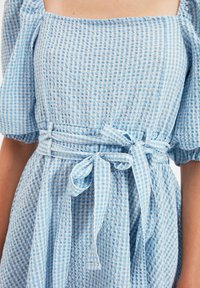 Světle modré kostkované šaty vyrobené z texturované látky, s nafouknutými rukávy, přiléhavým korzetem a opaskem na zavázání pro přidaný detail.