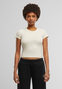Crop top crème à manches courtes en tissu doux avec un design ajusté et un logo imprimé subtil sur la poitrine, associé à un pantalon de jogging noir.