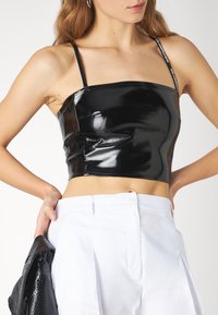 Top noir court en matériau brillant avec des fines bretelles, offrant un design ajusté et des coutures minimales, associé à un pantalon blanc.