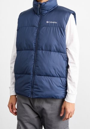 Person trägt eine ärmellose, blaue, gesteppte Columbia-Weste über einem weißen Langarmhemd und grauen Hosen, schaut nach vorne.