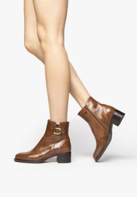 Braune Leder-Stiefeletten mit einem klobigen Absatz, ausgestattet mit einem seitlichen Reißverschluss und einer dekorativen Schnalle am Knöchel. Glatte Textur und schlanke Form.