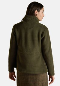 Žena videna zozadu, oblečená v olivovo zelenej fleecovej bunde s výšivkou "SOULES" na golieri a vzorovanou sukňou.