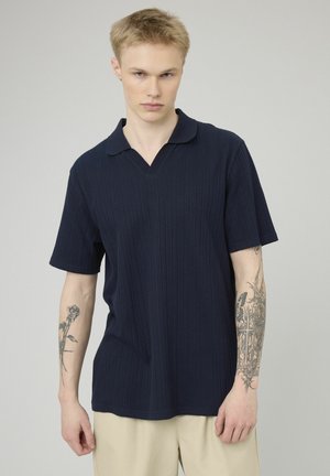 Jeune homme avec des tatouages sur les deux avant-bras, portant un polo à manches courtes bleu marine et un pantalon beige, sur un fond uni.