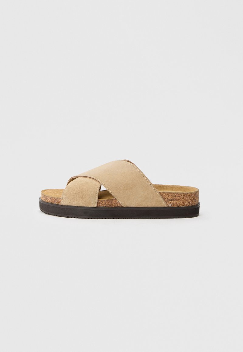 Sandale à glissière en daim beige avec un design à lanières croisées, semelle intermédiaire en liège et semelle extérieure en caoutchouc noir. Texture lisse et apparence minimaliste.