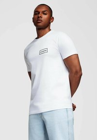 Ung mand iført en hvid T-shirt med korte ærmer, der har "OMBRE" logoet, og lyseblå jeans, stående mod en ensfarvet lys baggrund.