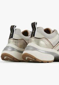 Sneakers hebben een witte bovenkant met metallic gouden accenten, een getextureerd mesh-gedeelte en een dikke rubberen zool. De hiel heeft een trekkoordje.