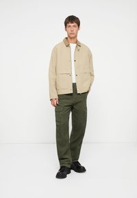 Beige cotton jacket with a brown collar, paired with green corduroy cargo pants featuring multiple pockets. Black shoes complete the outfit.