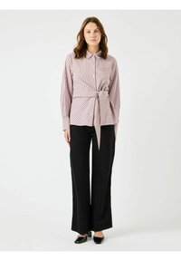 Gestreepte button-up shirt in lichtroze en wit, geknoopt in de taille, gecombineerd met zwarte high-waisted broek met wijde pijpen en zwarte schoenen.