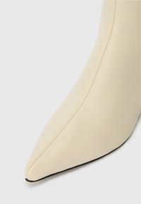 Botte de cheville beige à bout pointu avec une finition lisse et élégante et une semelle noire. Présente un détail de couture épuré sur le côté.