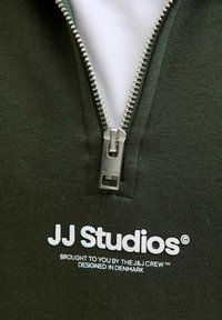 Grön zip-up sweatshirt med en silverdragkedja, vit innervägg och präglad "JJ Studios"-logga med tilläggstext i vitt.