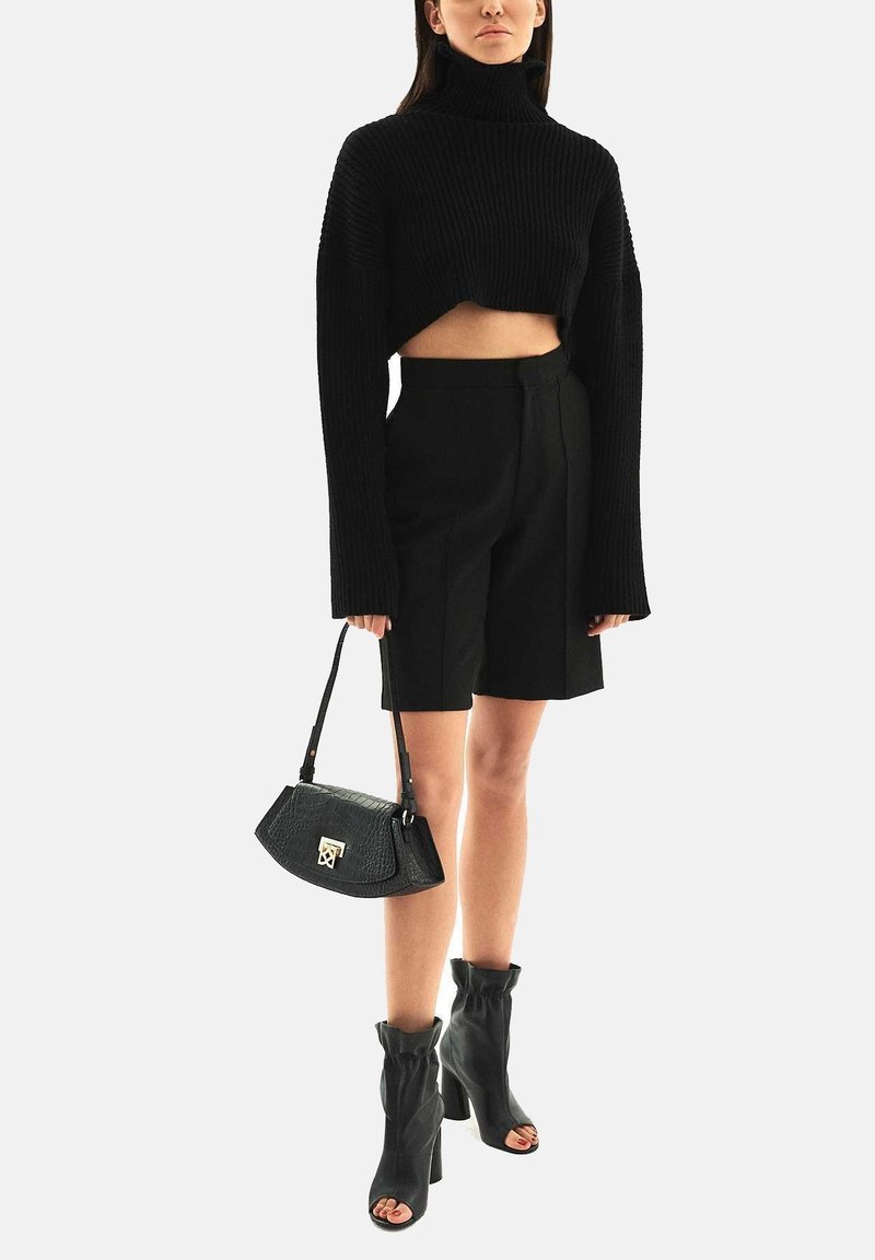 Femme portant un pull court noir à col roulé, un short taille haute jusqu'au genou, des bottines noires à talons ouverts, tenant un sac à main noir.
