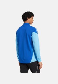 Blaues Sportjackett mit langen Ärmeln, das an den Ärmeln einen hellblauen Akzent aufweist, einen Reißverschlusskragen hat und ein kontrastierendes Panele-Design besitzt.