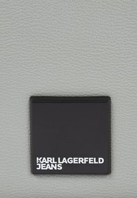 Pelle grigia testurizzata con un'etichetta rettangolare nera in rilievo con la scritta "KARL LAGERFELD JEANS" in bianco, posizionata al centro.