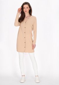 Cardigan lungo beige con scollatura a V, chiusura con bottoni e maniche lunghe. Indossato sopra pantaloni bianchi a vestibilità aderente e sneakers bianche. Texture in cotone morbido.