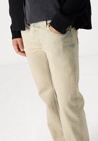 Vaqueros de mezclilla beige con corte recto, ligero desgaste, diseño de cinco bolsillos y detalles de costura visibles, llevados con un cárdigan negro.