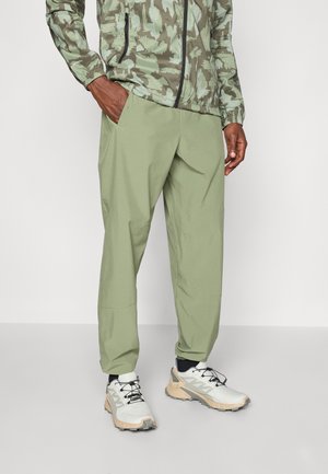 Lekkie, oliwkowo-zielone spodnie z elastycznymi mankietami i bocznymi kieszeniami, zestawione z białymi i beżowymi sportowymi butami o fakturowanej podeszwie.