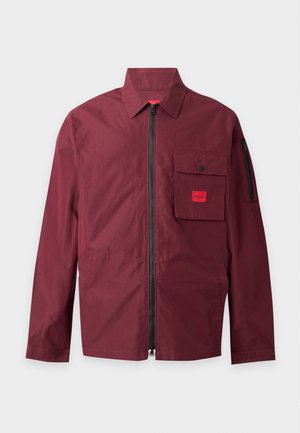 Homme portant une veste zippée bordeaux avec poche avant, pantalon cargo noir et baskets foncées, debout devant un fond uni.