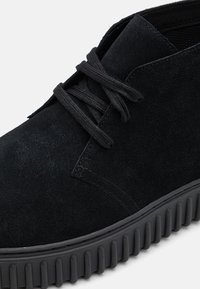 Scarpa in suede nera con punta arrotondata, tomaia testurizzata e lacci neri piatti. Presenta una suola in gomma spessa e rigata per una maggiore trazione.