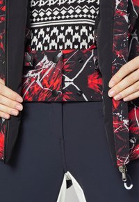 Svart och röd jacka med abstrakt målarliknande mönster, dubbla tryckknappar och hög hals, bärs över en mönstrad tröja och marinblå byxor.