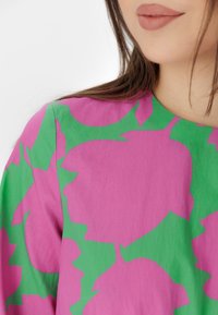 Woman wearing a pink and green floral patterned top with long brown hair and nude lipstick, shown from the neck to shoulders.