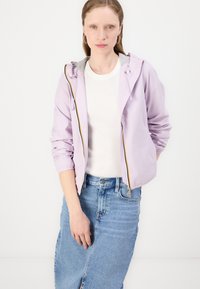 Giacca a zip viola chiaro con cappuccio, dotata di una fodera grigia e di una cerniera con accento giallo. Indossata sopra una maglia bianca e una gonna di jeans blu.