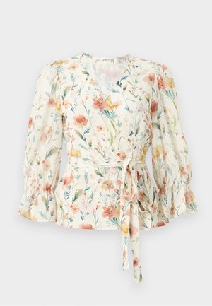 Femme portant un chemisier cache-cœur floral crème avec des manches volantées et un pantalon crème taille haute à jambes larges, se tenant devant un fond uni.