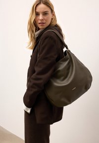 Olíva zöld bőr kézitáska laza fazonnal, arany logó részlettel és egyetlen pánttal, a vállon viselve, semleges háttér előtt.