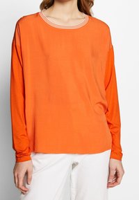 Frau in einem langärmeligen orangen Oberteil mit gestreiftem Rundhalsausschnitt und weißen Hosen, dargestellt von den Schultern bis zur Hüfte.