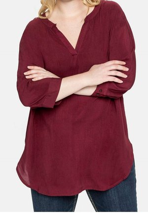 Kobieta w bordowej bluzce z długim rękawem i dekoltem w serek oraz ciemnoniebieskich dżinsach, z rękami skrzyżowanymi na piersi.