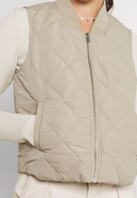 Beżowa pikowana kamizelka z zamkiem błyskawicznym z przodu, kieszeniami i ściągaczem na kołnierzu. Materiał ma gładką teksturę i subtelny połysk.