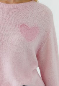 Pull rose tendre avec un détail en tricot coeur rose plus foncé sur la partie supérieure de la poitrine, porté par une personne sur un fond uni.