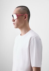 Rózsaszín műanyag keretes napszemüveg nagy, tükörlencsékkel. Egy fehér pólót viselő személyen, amelyen diszkrét vállrészletek találhatók.