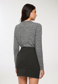 Donna con lunghi capelli scuri che indossa una maglietta grigia a maniche lunghe e una mini gonna nera a righe, rivolta di lato verso uno sfondo semplice.