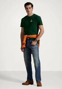 En ung man som bär en mörkgrön polo-tshirt, blå jeans, bruna skor och en orange tröja knuten runt midjan.