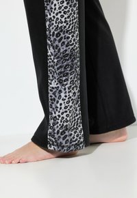 Bosé nohy obuté v černých kalhotách se svislým šedým a černým leopardím potiskem na bocích na bílém pozadí.
