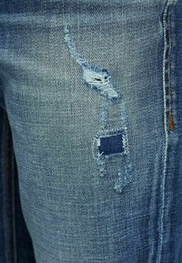 Worn blue denim fabric with two distressed patches, one revealing dark blue material underneath, and visible stitching along the seam.