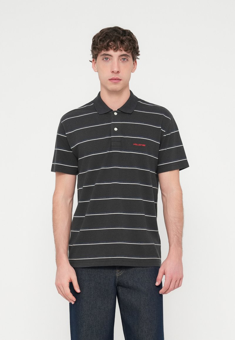 Σκούρο γκρι πόλο μπλουζάκι με λευκές και ανοιχτό μπλε ρίγες, πατιλέτα με τρία κουμπιά και κόκκινο λογότυπο "Hollister" στο στήθος.