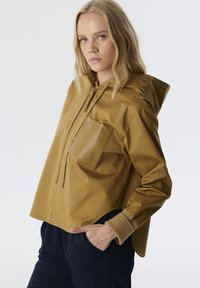 Bruine hooded jas met een unieke voorkantzak, met een gladde textuur en een relaxed fit. De mouwen zijn ribbelig en er is een extra zak aanwezig.