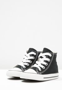 Svart kanvas høyhælte sneakers med hvite snører, gummitupper og kontrasterende hvite sømmer. Flate såler med svarte striper på bunnen.